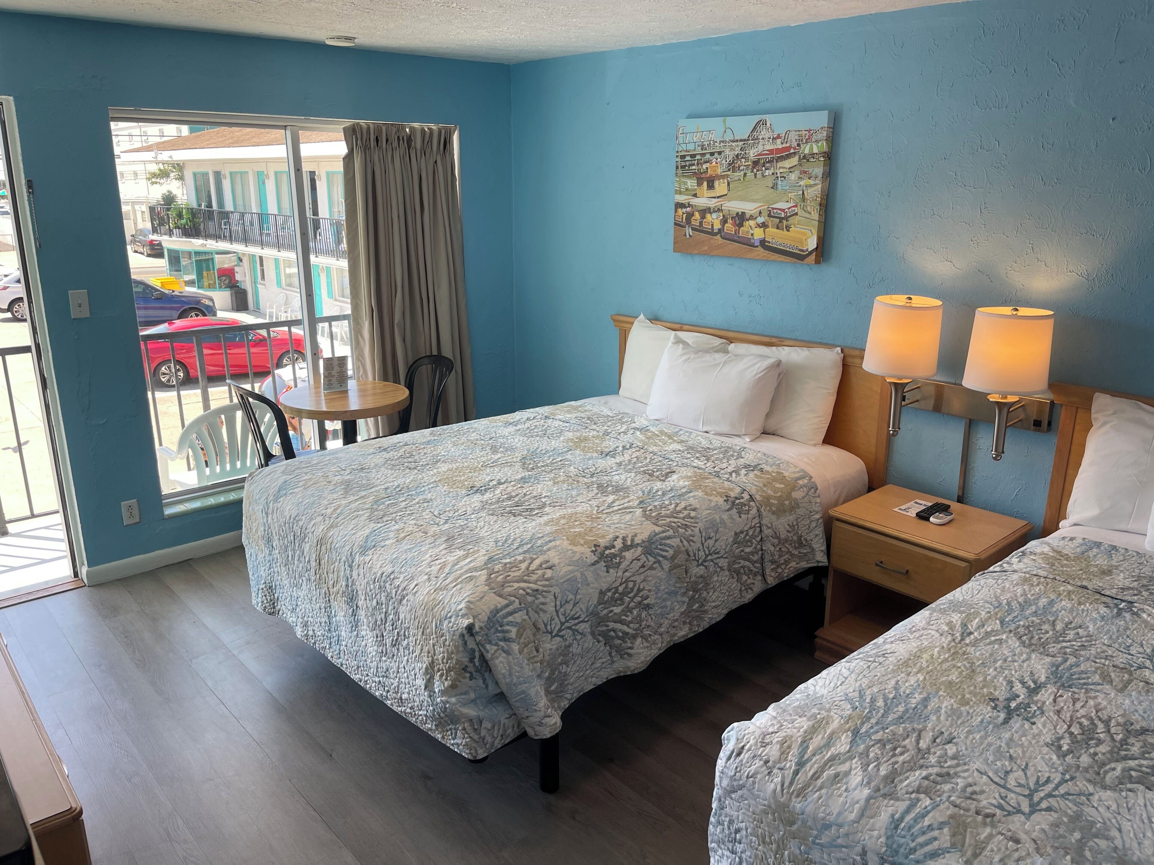 A hotel room with two beds, a window with a view of a street, a painting on the wall, and a nightstand with a lamp.