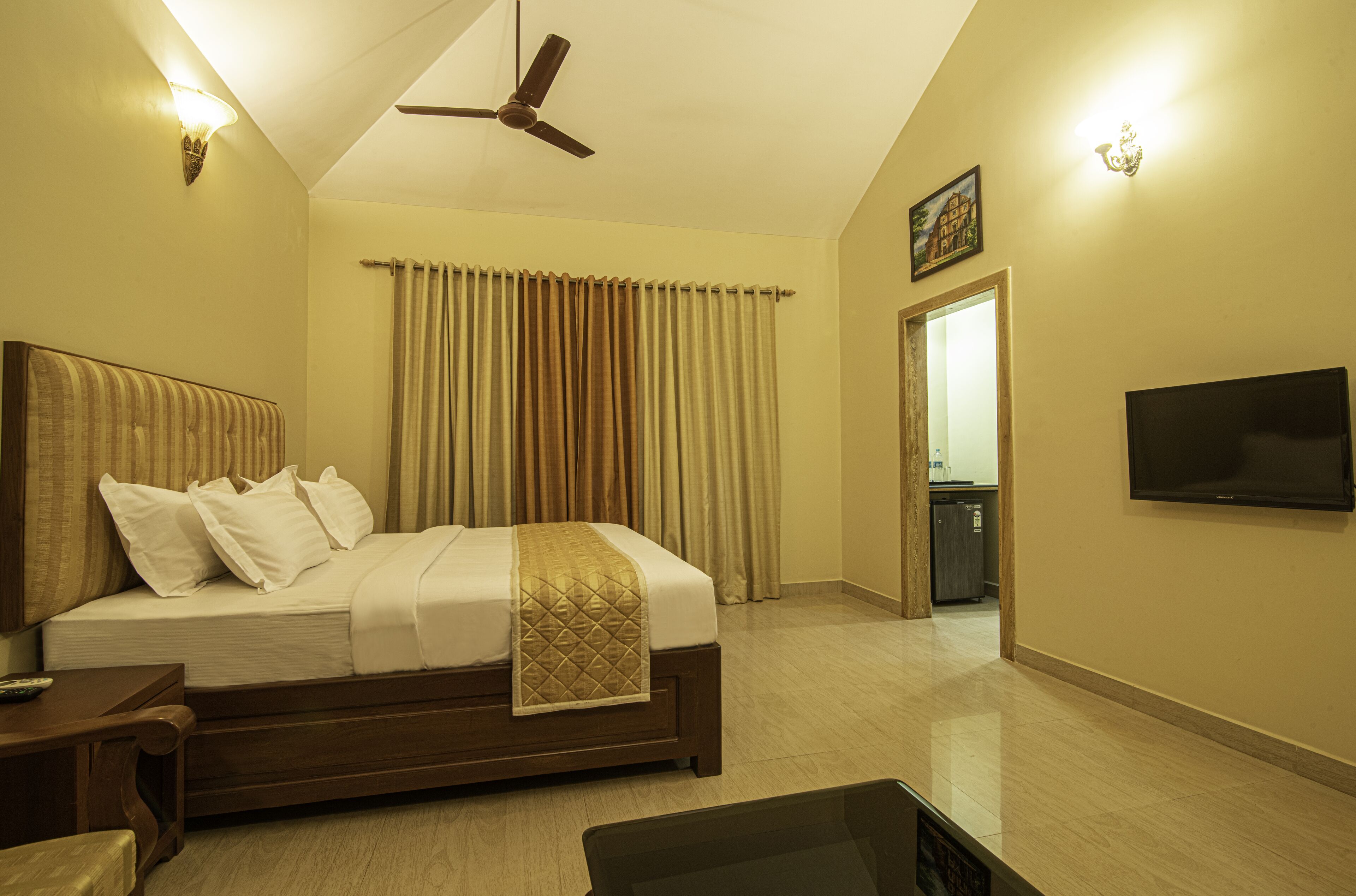 Habitación de hotel con cama, televisión, ventilador de techo y vista al baño.