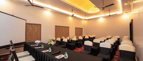 Un salón de conferencias con una larga mesa, sillas, una pantalla de proyector y ventiladores de techo.