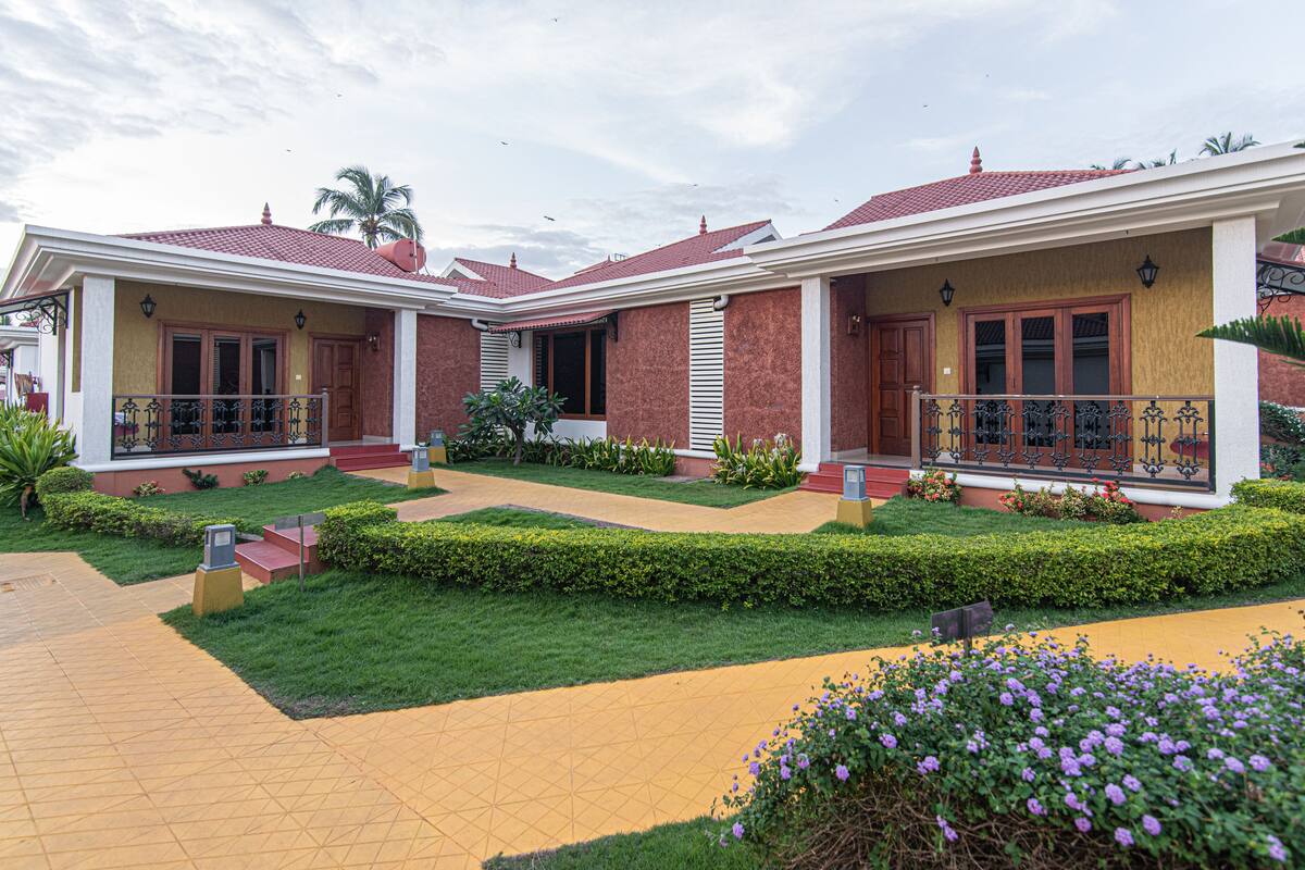Una casa de dos pisos con techo de tejas rojas, un porche con barandilla y un jardín bien cuidado con setos y flores.