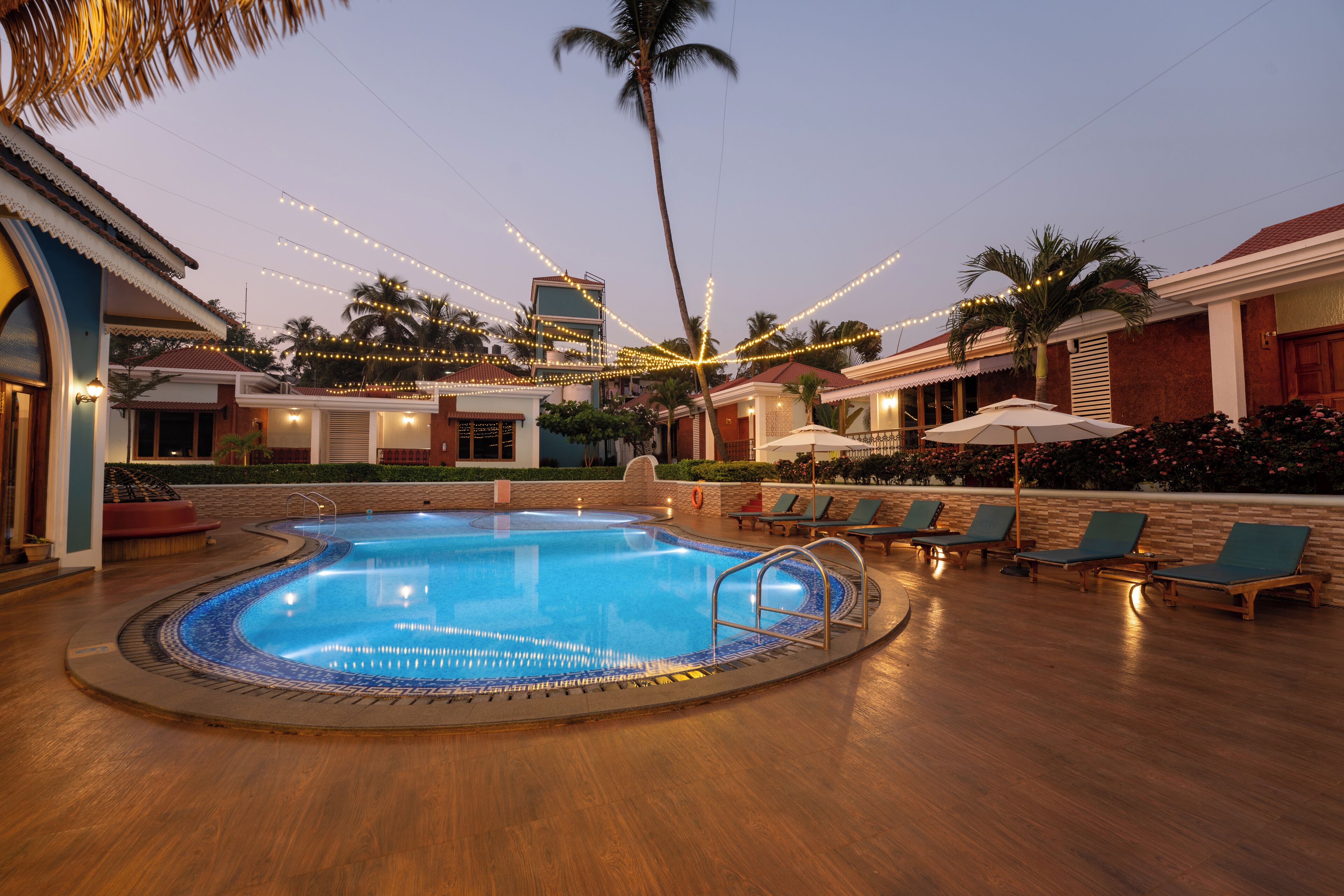 A swimming pool with a tiled edge, surrounded by lounge chairs and umbrellas, with string lights overhead.