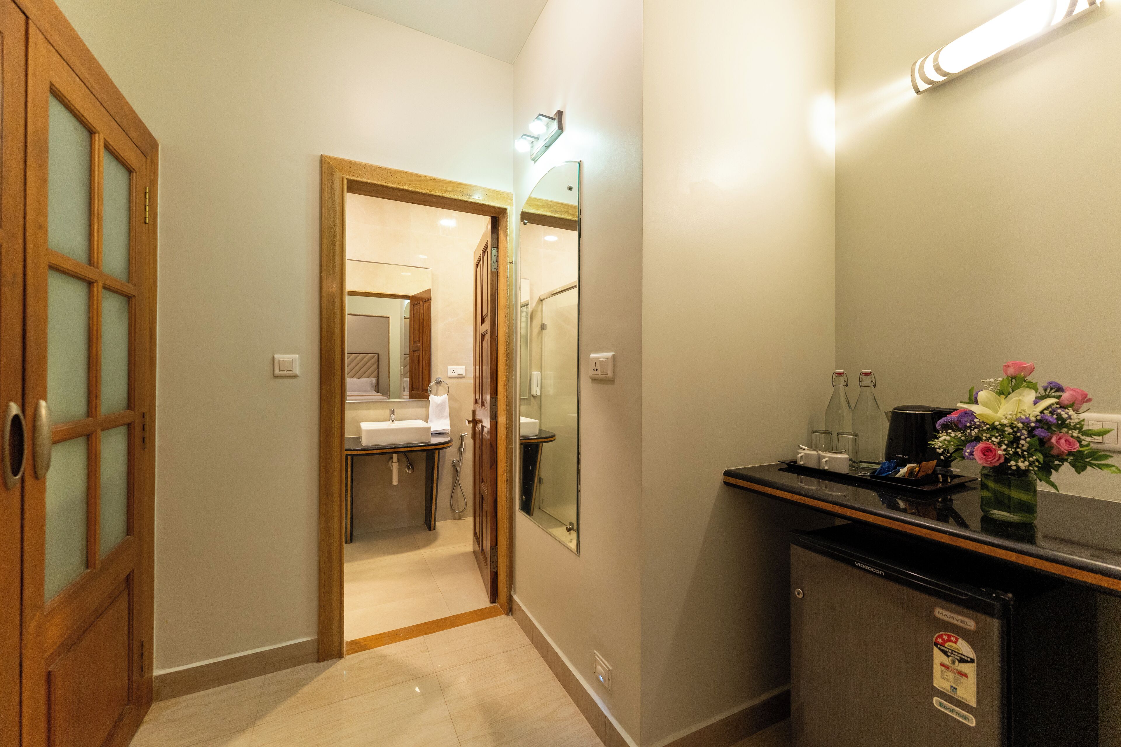 Un pasillo de hotel con una puerta de madera, un espejo, un refrigerador y un jarrón de flores.