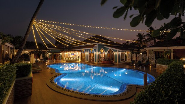 Una piscina de borde curvo, rodeada de sillas de descanso y sombrillas, con luces colgantes.