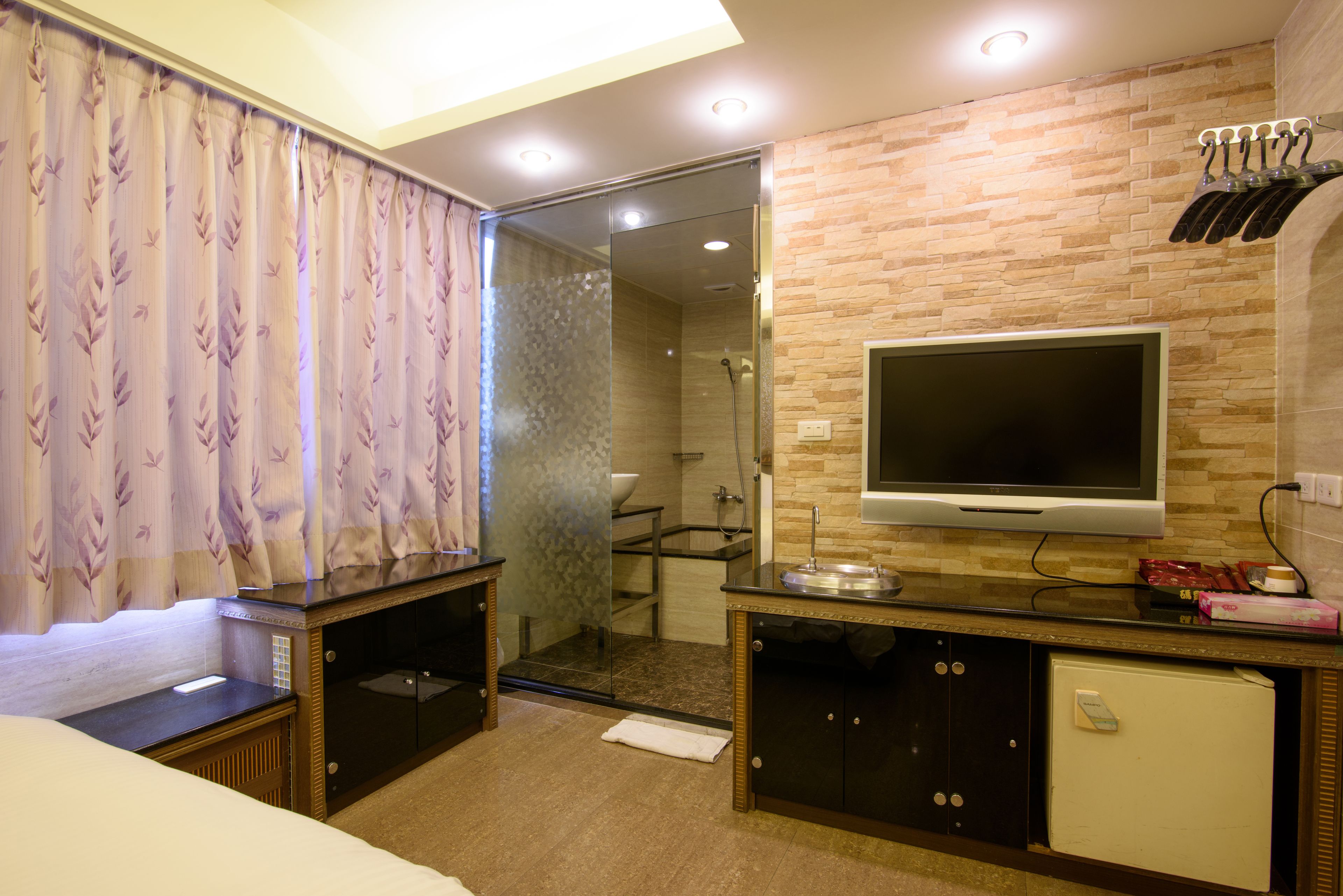 A hotel room with a bed, a TV mounted on the wall, a shower area, and a small refrigerator.