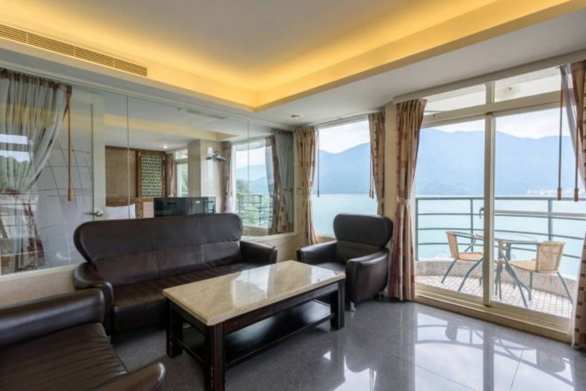 A modern hotel room with a sofa, armchair, and a small table. It features large windows with a view of the sea and mountains.