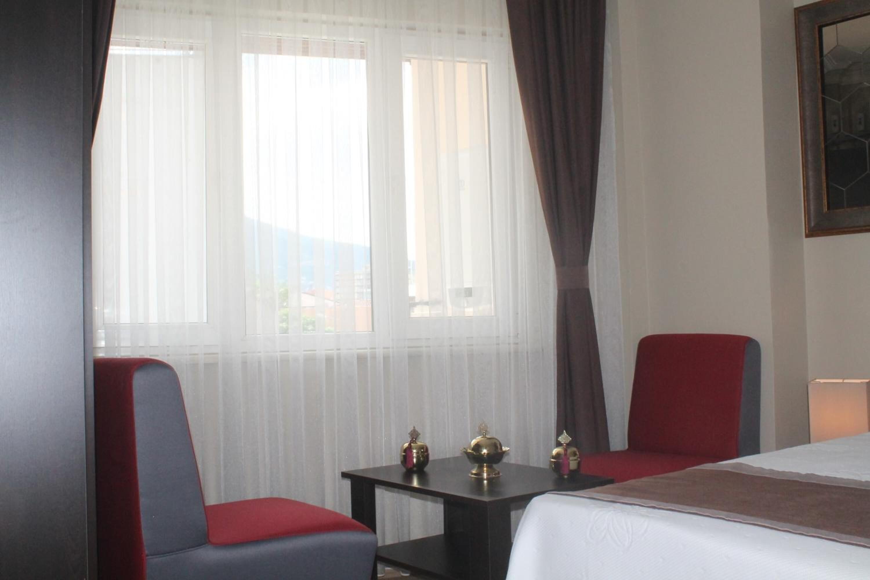 A hotel room with a bed, two red chairs, a small table with decorative items, and a window with sheer curtains.