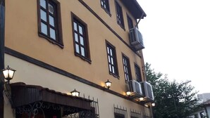 A two-story building with a beige facade, multiple windows with dark frames, and a wooden door. There are outdoor lights and a hanging flower arrangement near the entrance. - Lena Central Flats (Bursa)