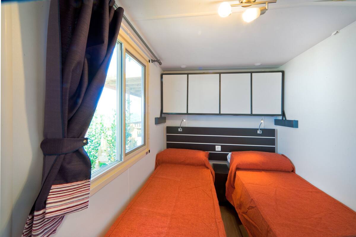 A small, compact room with two single beds, a window with curtains, a headboard with storage, and a wall-mounted light fixture.