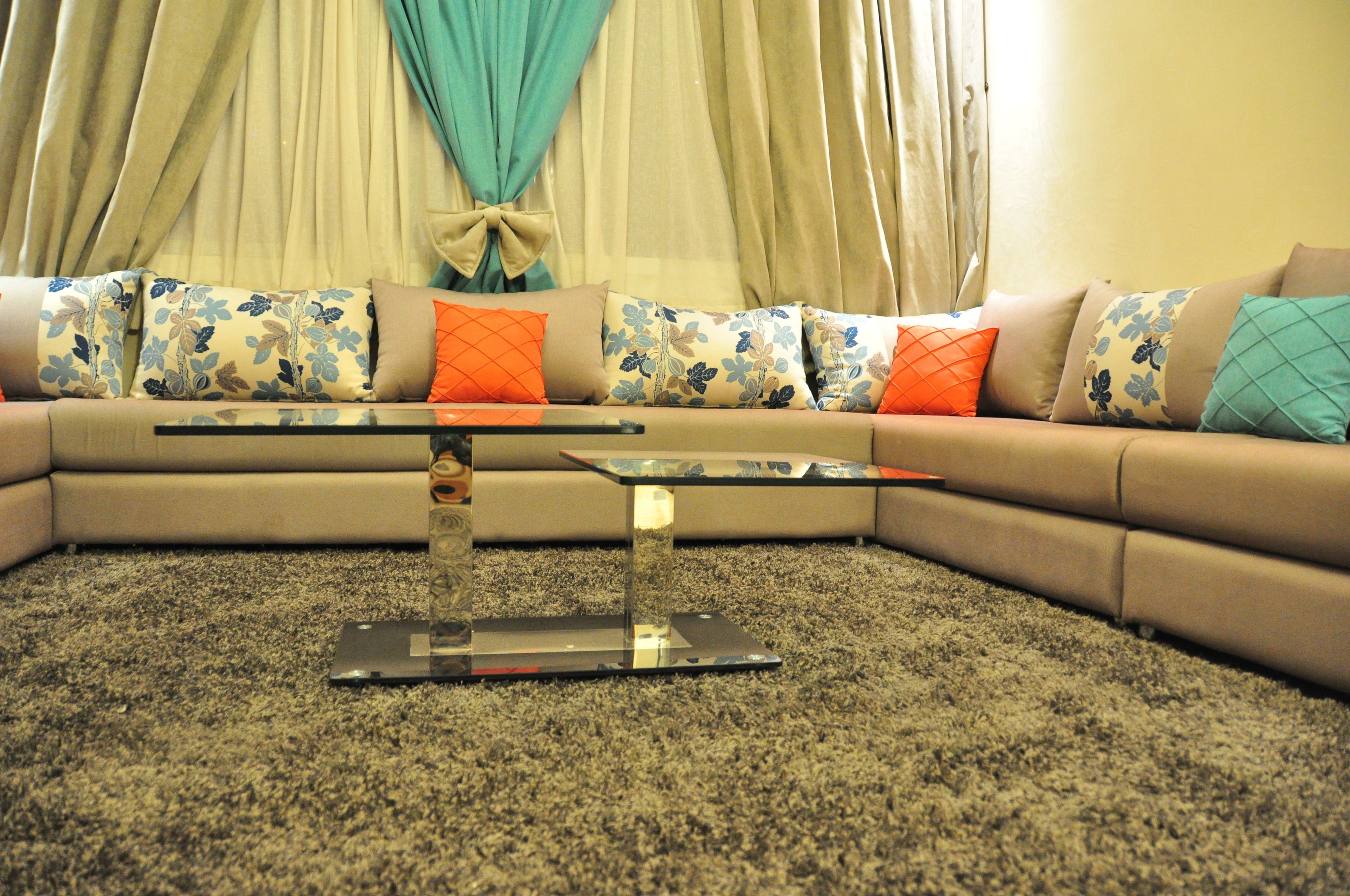 A living room with a beige sofa, floral cushions, a glass coffee table, and a carpeted floor.