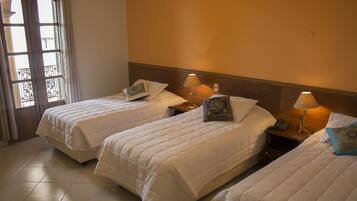 Quarto de hotel com três camas de solteiro, cada uma com travesseiro e mesa de cabeceira com abajur.