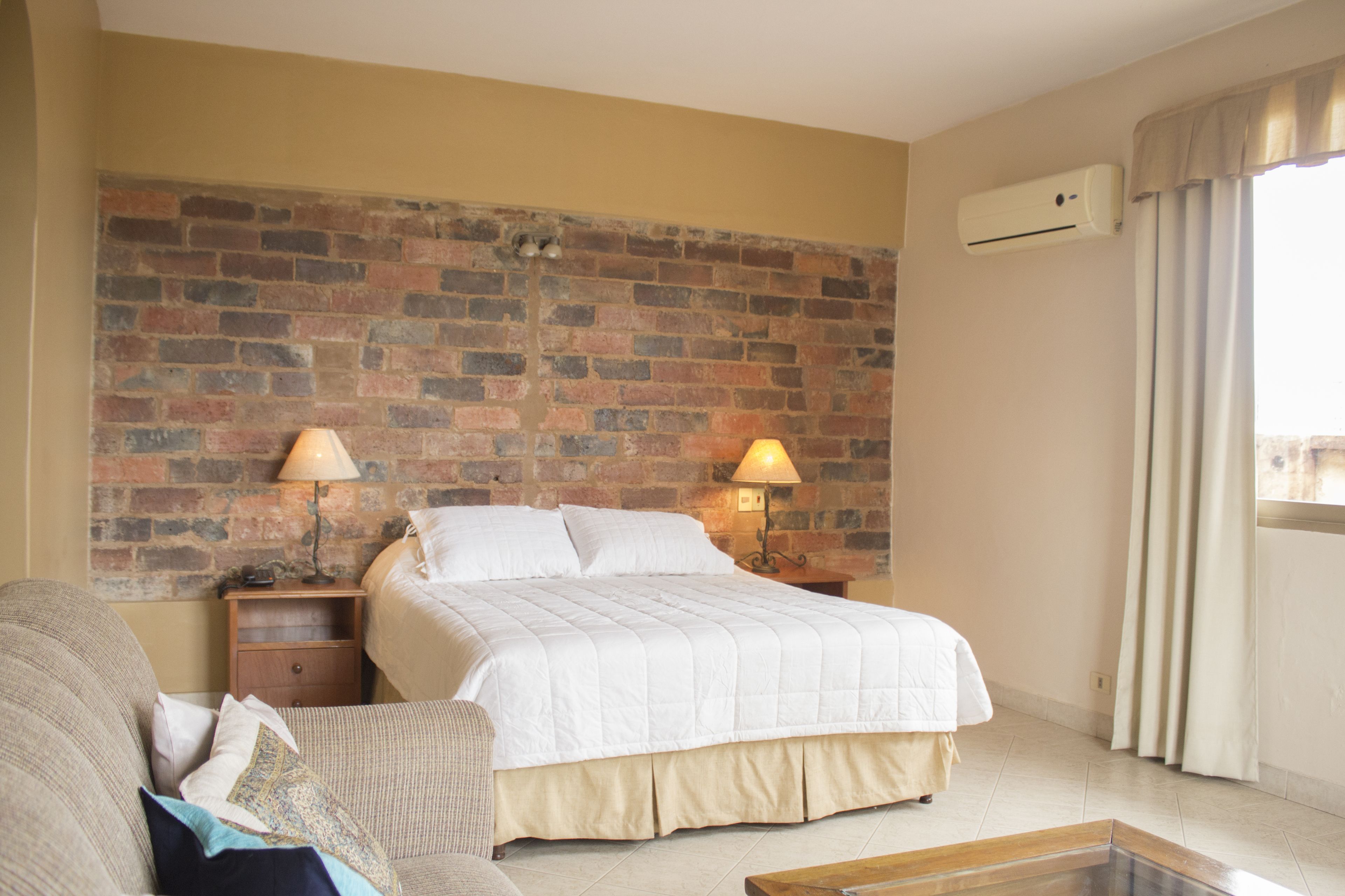 Habitación de hotel con pared de ladrillo, cama con ropa blanca, dos lámparas de noche, mesita de noche de madera, sofá color beige y ventana con cortinas.