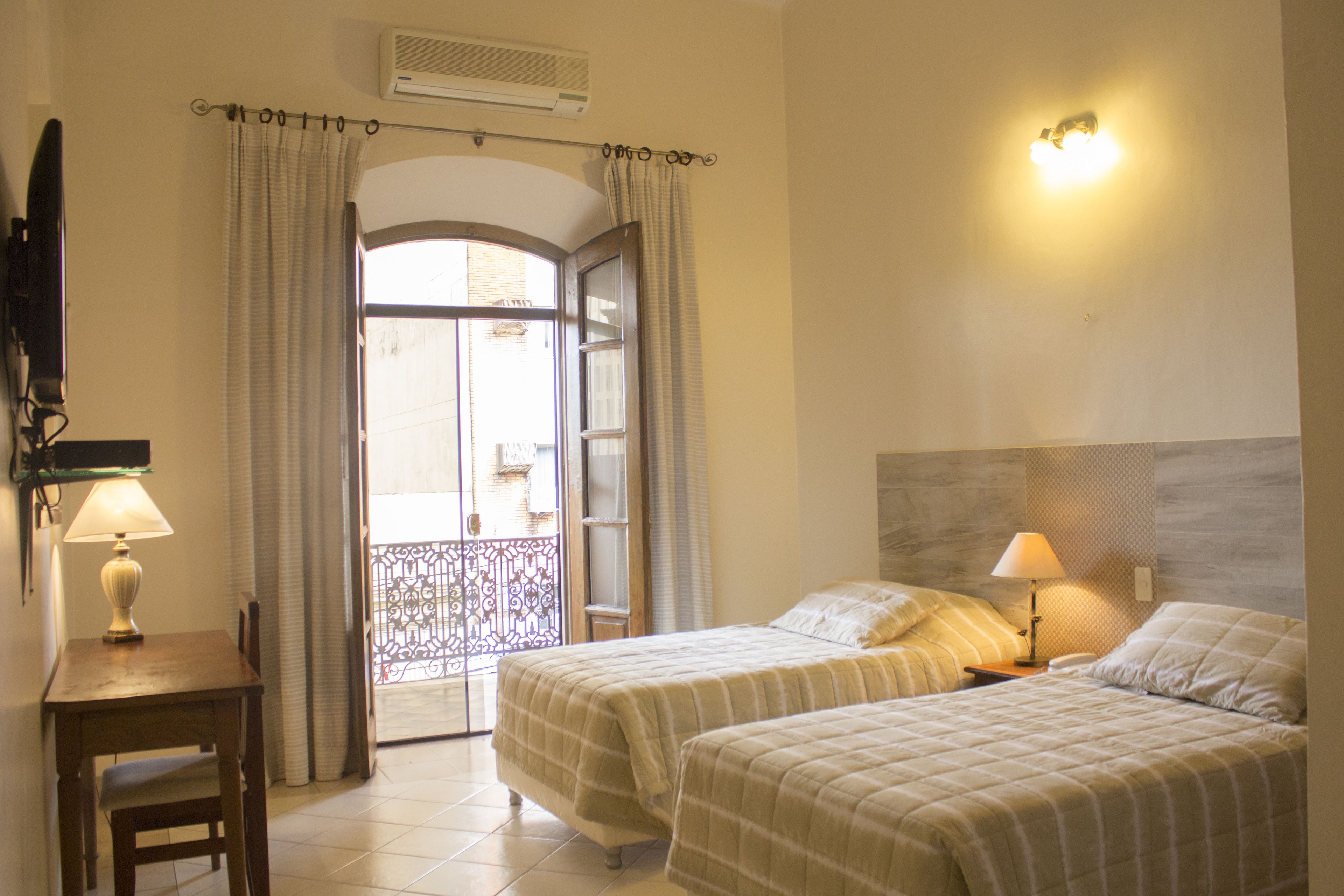 A hotel room with two beds, a desk with a lamp, a chair, a balcony with a view, and a ceiling-mounted air conditioner.