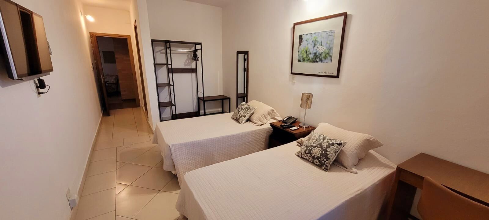 Ein Hotelzimmer mit zwei Betten, einem Kleiderschrank, einem Schreibtisch, einem Stuhl, einem Fernseher und einem gerahmten Bild an der Wand.