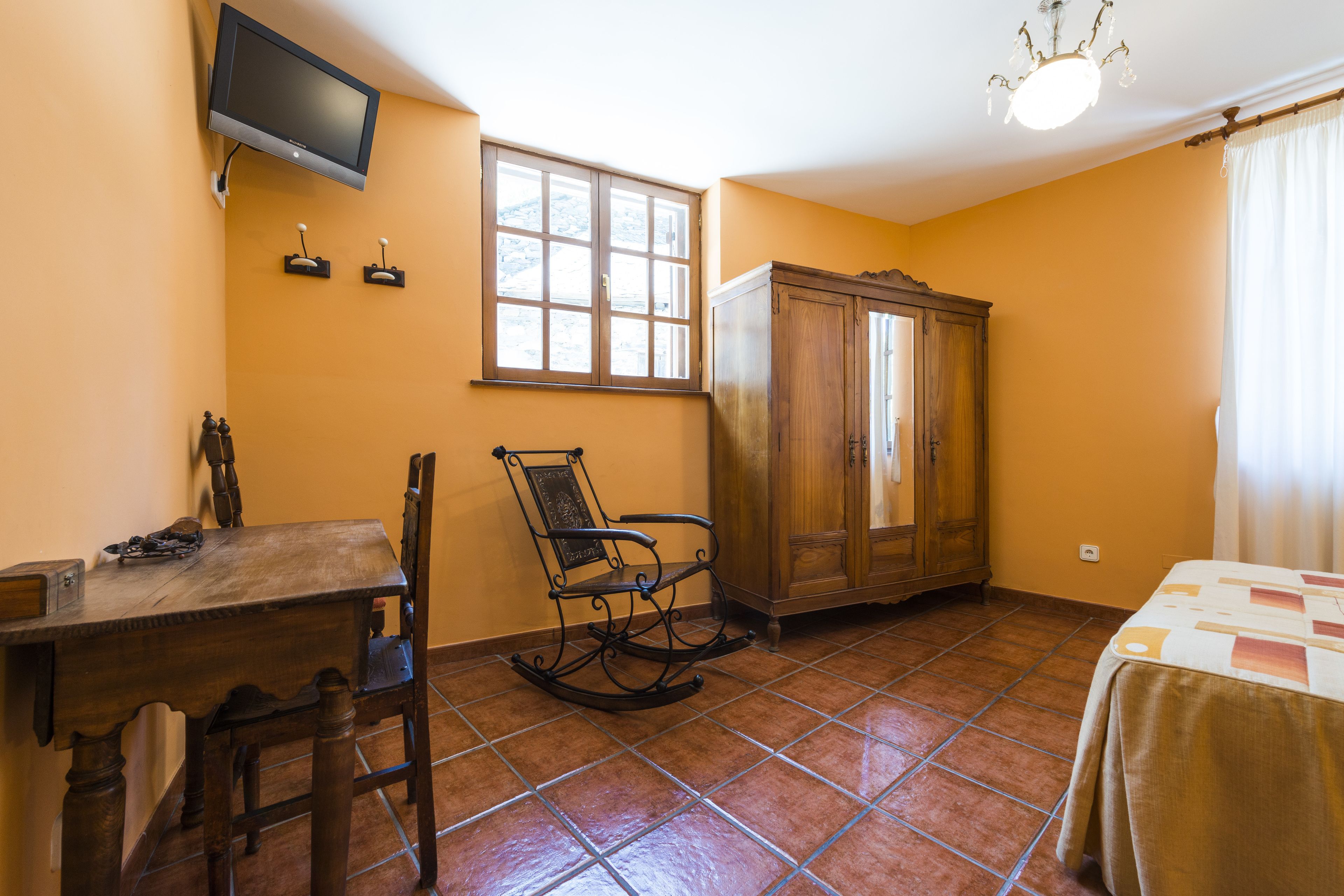 A room with a wooden table, a rocking chair, a cabinet, a television mounted on the wall, and a window with curtains.