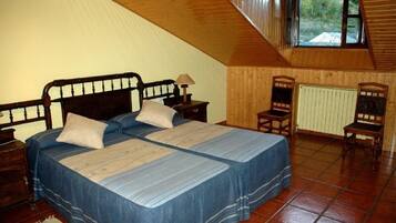 A bedroom with a wooden sloped ceiling, two beds with blue bedding, a nightstand, a chair, and a window with a view of trees.