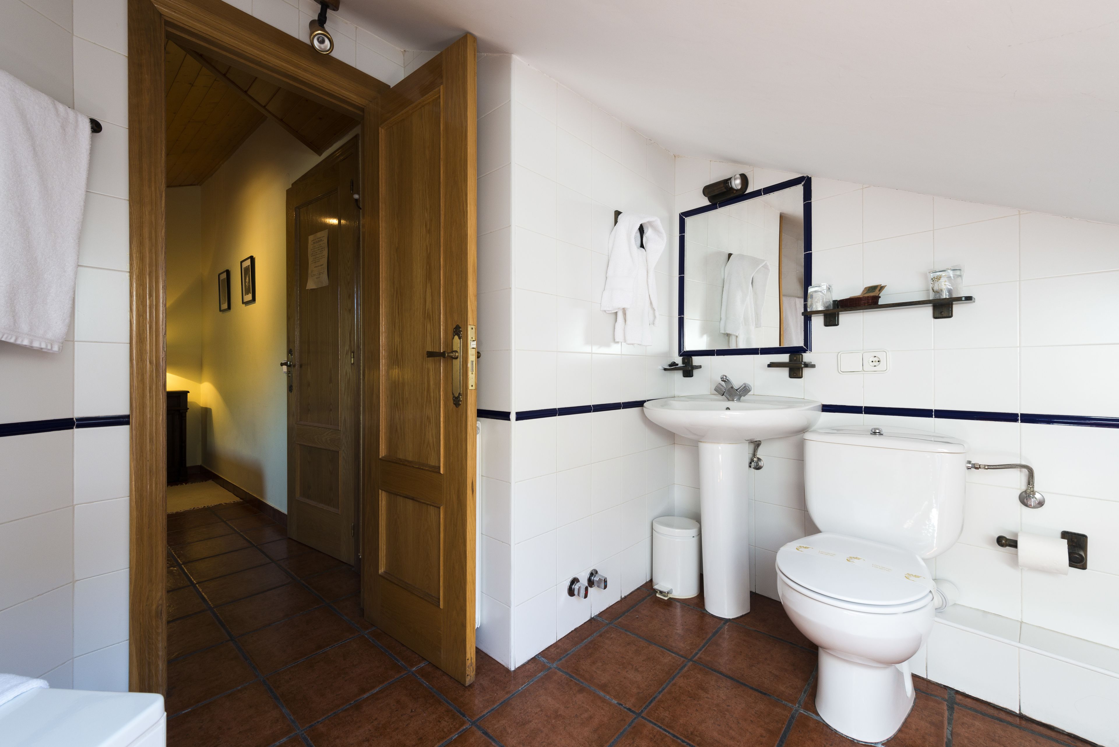Un baño con puerta de madera, azulejos blancos, un lavamanos, un inodoro y un espejo.