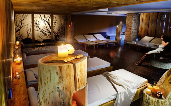 Un cuarto de spa con mobiliario de madera, una mesa de madera central con velas y una persona relajándose en una silla lounge.