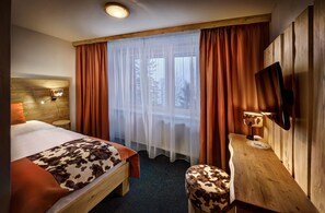 A hotel room with a bed, a wooden desk, a chair, a TV, and a window with curtains.