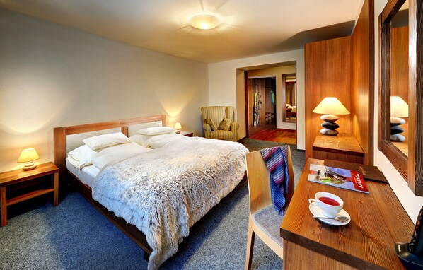 Una habitación de hotel con una cama grande, una mesita de noche de madera con lámpara, una silla, un escritorio de madera con lámpara y un espejo.