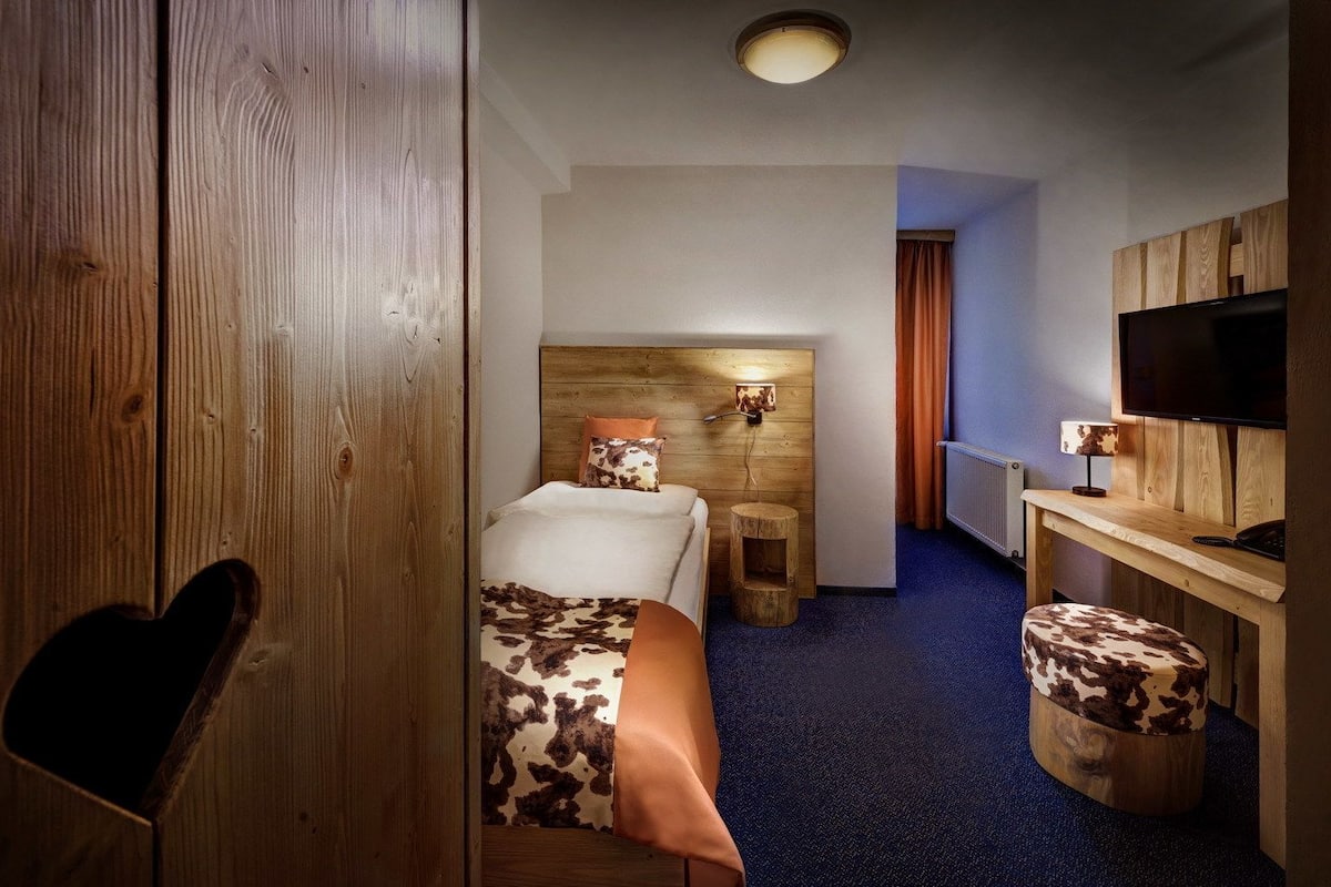 Hotelkamer met een houten hoofdeinde, een bed met een deken met een wit en bruin patroon, een klein houten nachtkastje met een lamp, een houten bankje en een televisie op een houten standaard.