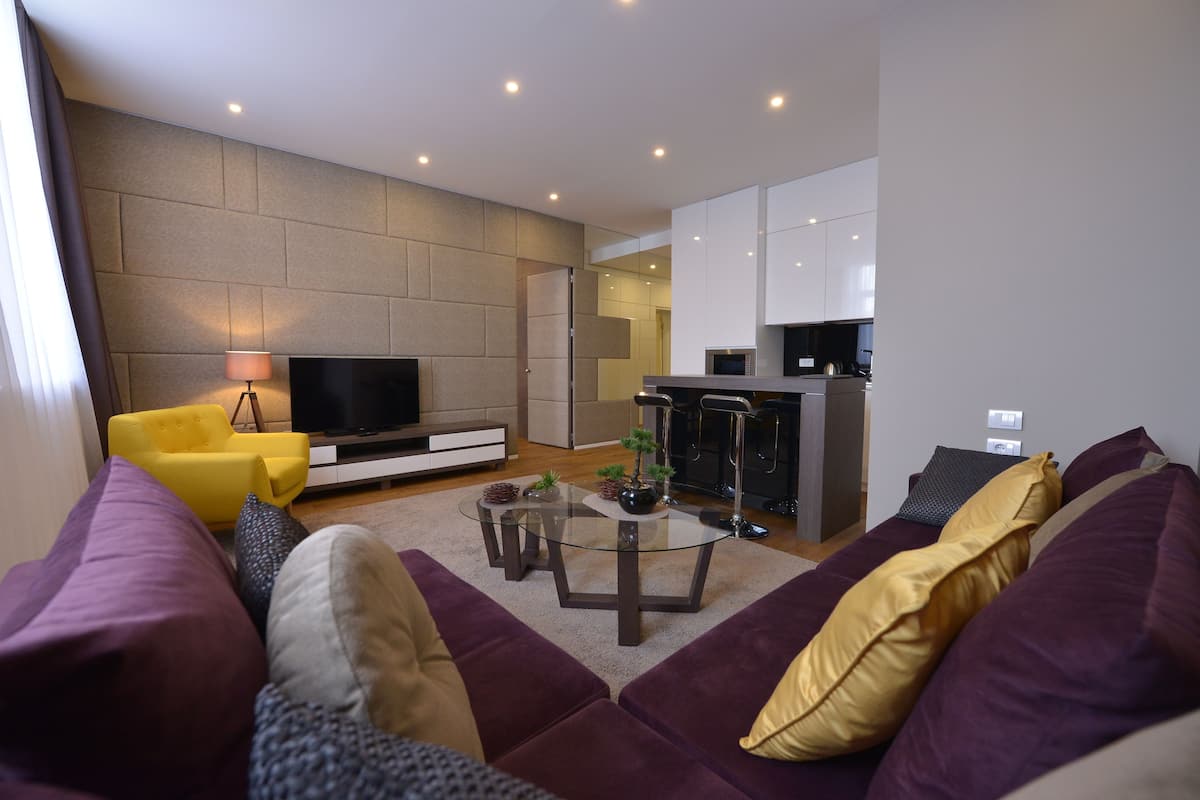 Un salon moderne avec un ensemble de canapés, une table basse, un téléviseur à écran plat et un coin cuisine à l’arrière-plan.