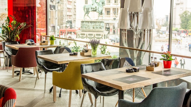 A modern dining area with multiple tables and chairs, large windows offering a view of the cityscape, and decorative plants. - Boutique Rooms (Belgrade)