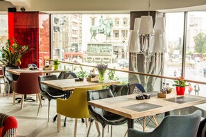 A modern dining area with multiple tables and chairs, large windows offering a view of the cityscape, and decorative plants.