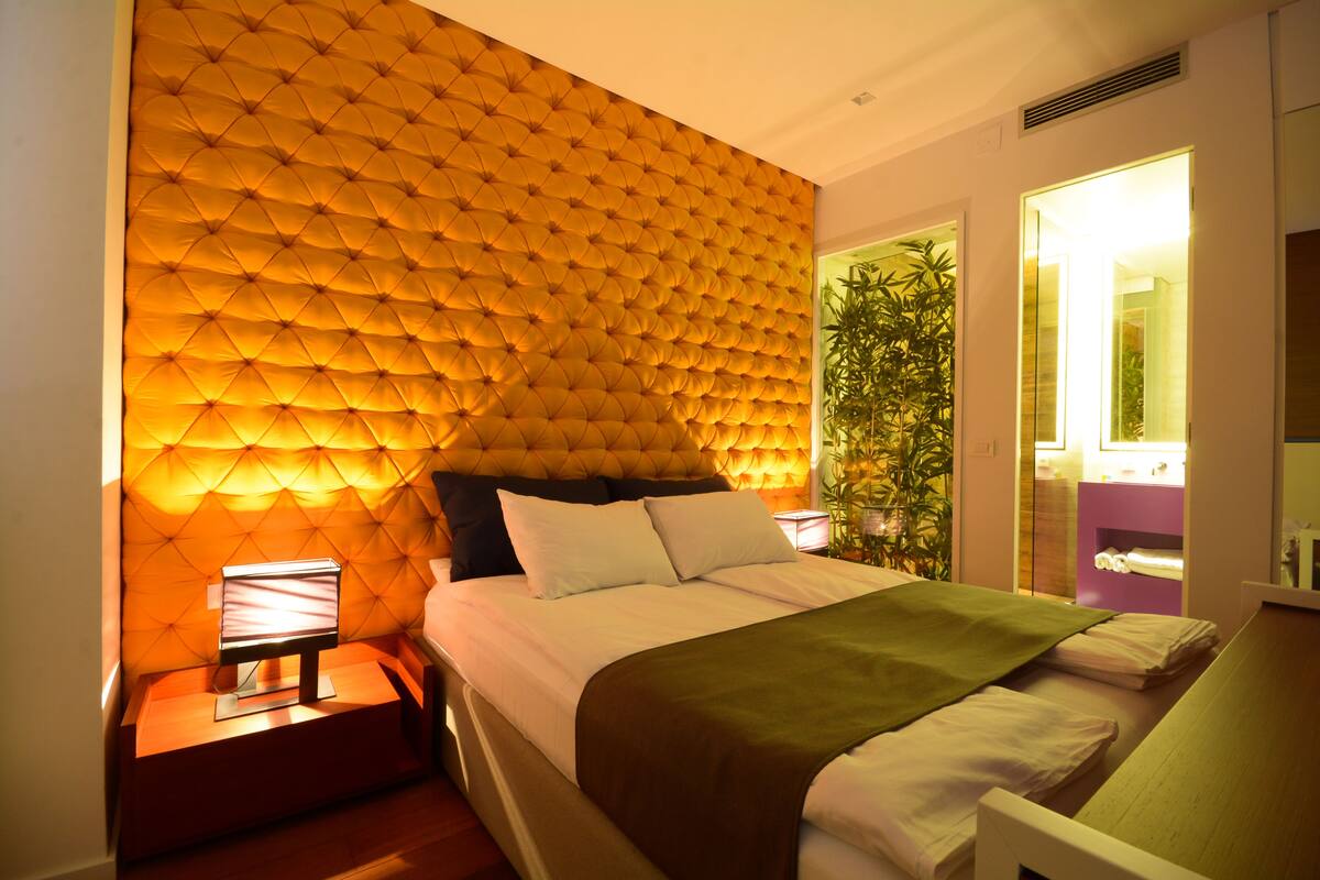 Une chambre à coucher moderne avec une tête de lit capitonnée, une table de chevet avec une lampe, un lit recouvert de draps blancs et un plaid vert.