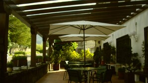 Un patio al aire libre con mesas y sillas bajo una pérgola, rodeado de plantas en macetas y una pared de piedra.