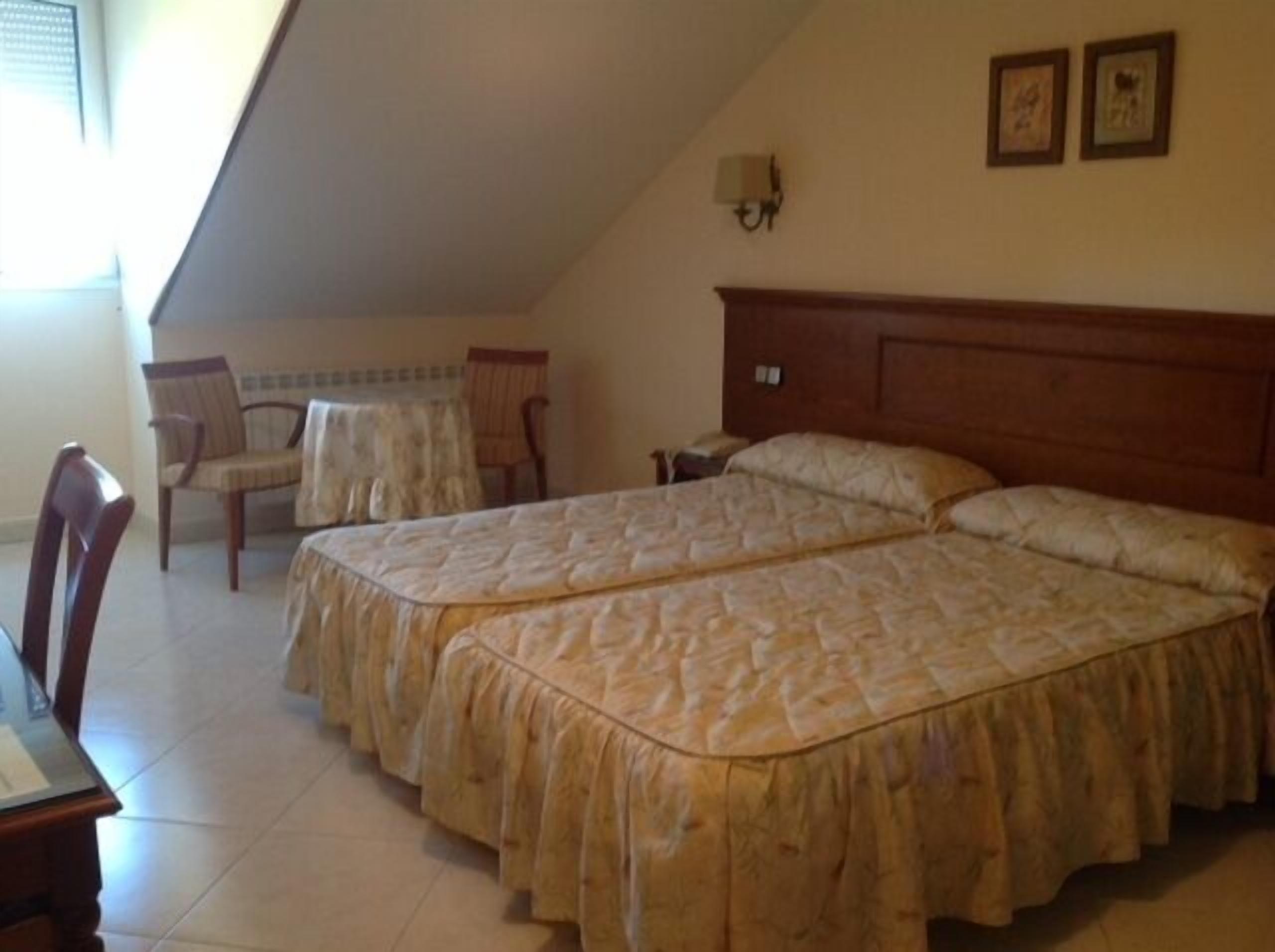 A bedroom with two beds, a chair, a small table, and a wall-mounted lamp.