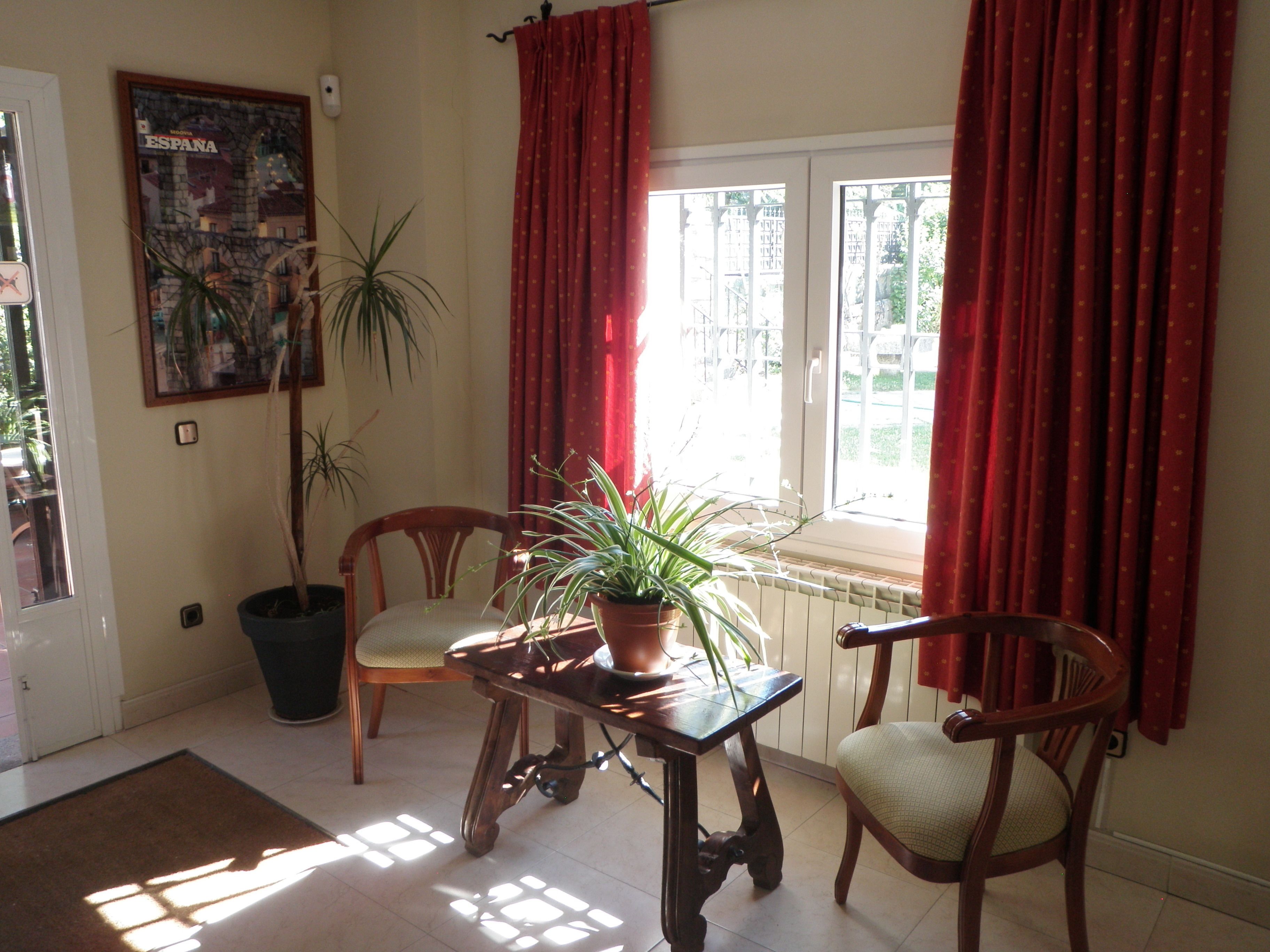 Una habitación con una mesa de madera, dos sillas, una planta en maceta, un cartel en la pared y una ventana con cortinas rojas.