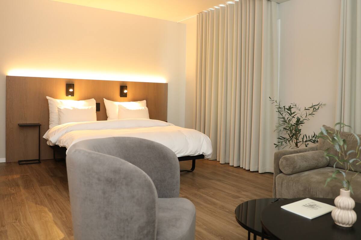 A hotel room with a bed, two bedside tables, a sofa, a small table with a vase, and a potted plant.