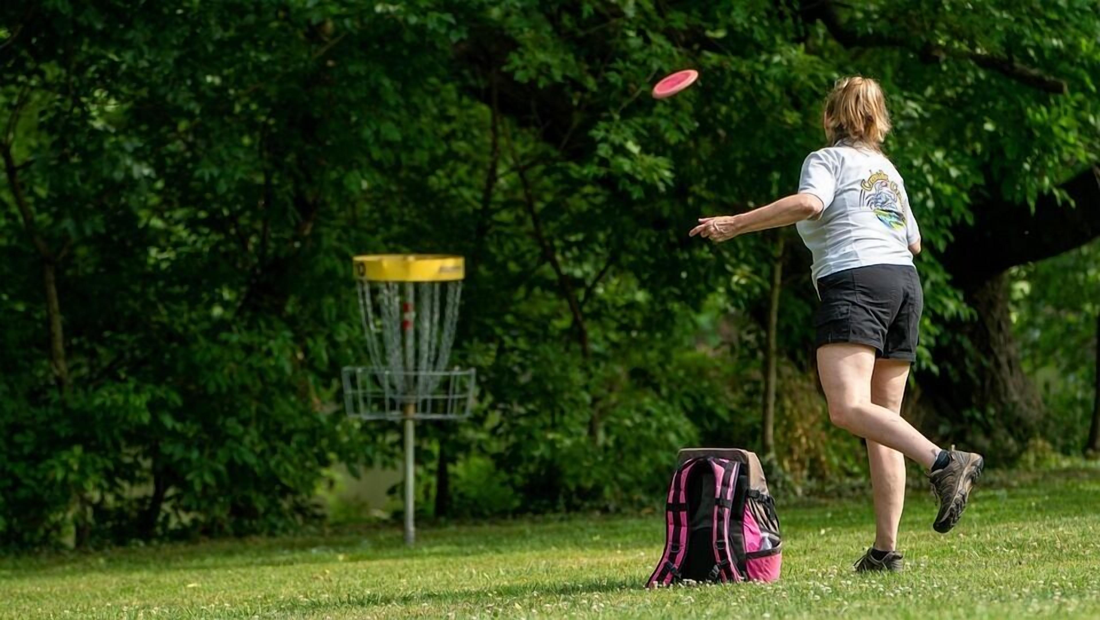 A person playing frisbee in a park with a disc golf basket and a backpack on the ground.