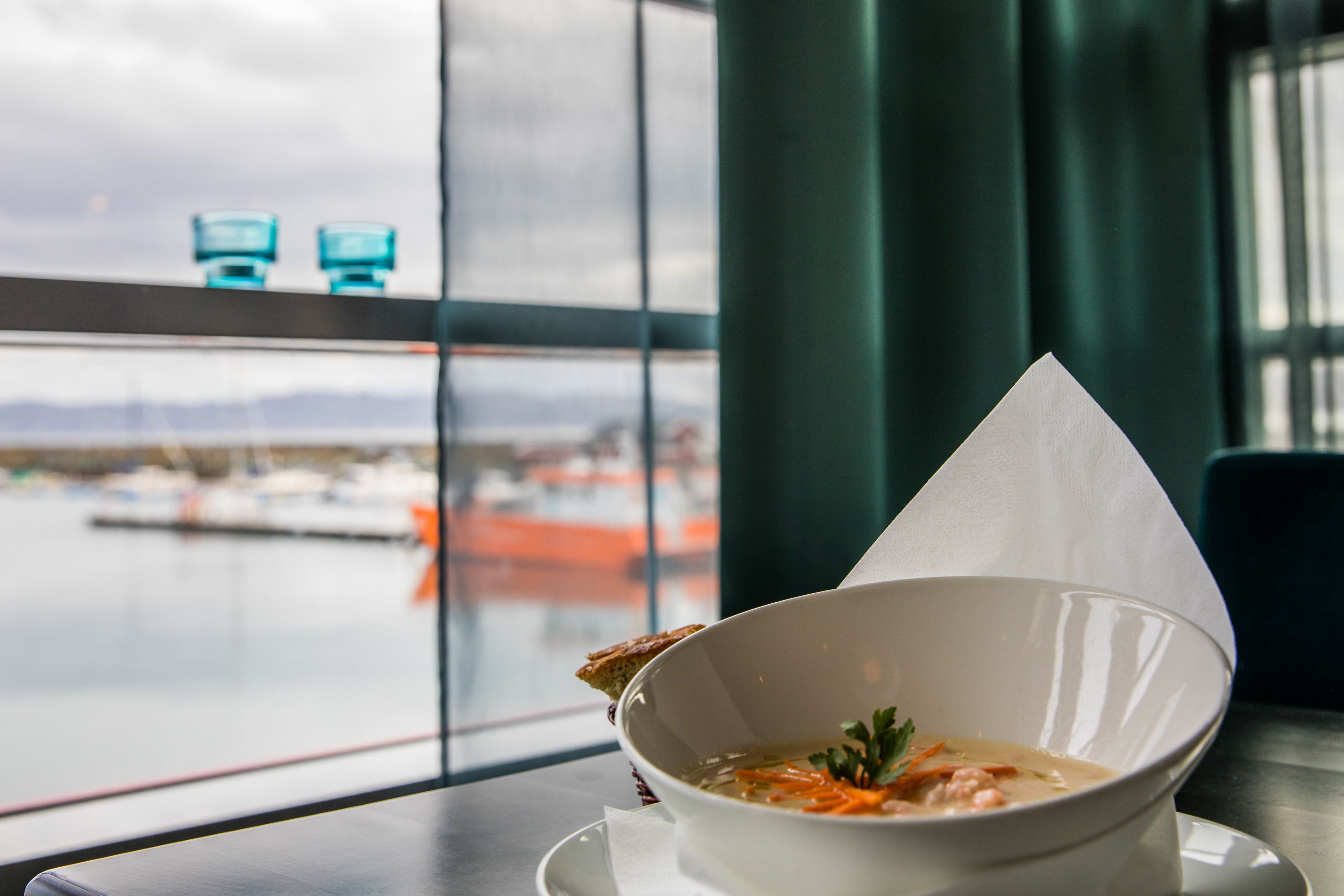 Un bol de soupe est posé sur une table, et de grandes fenêtres offrent une vue sur un port de plaisance.
