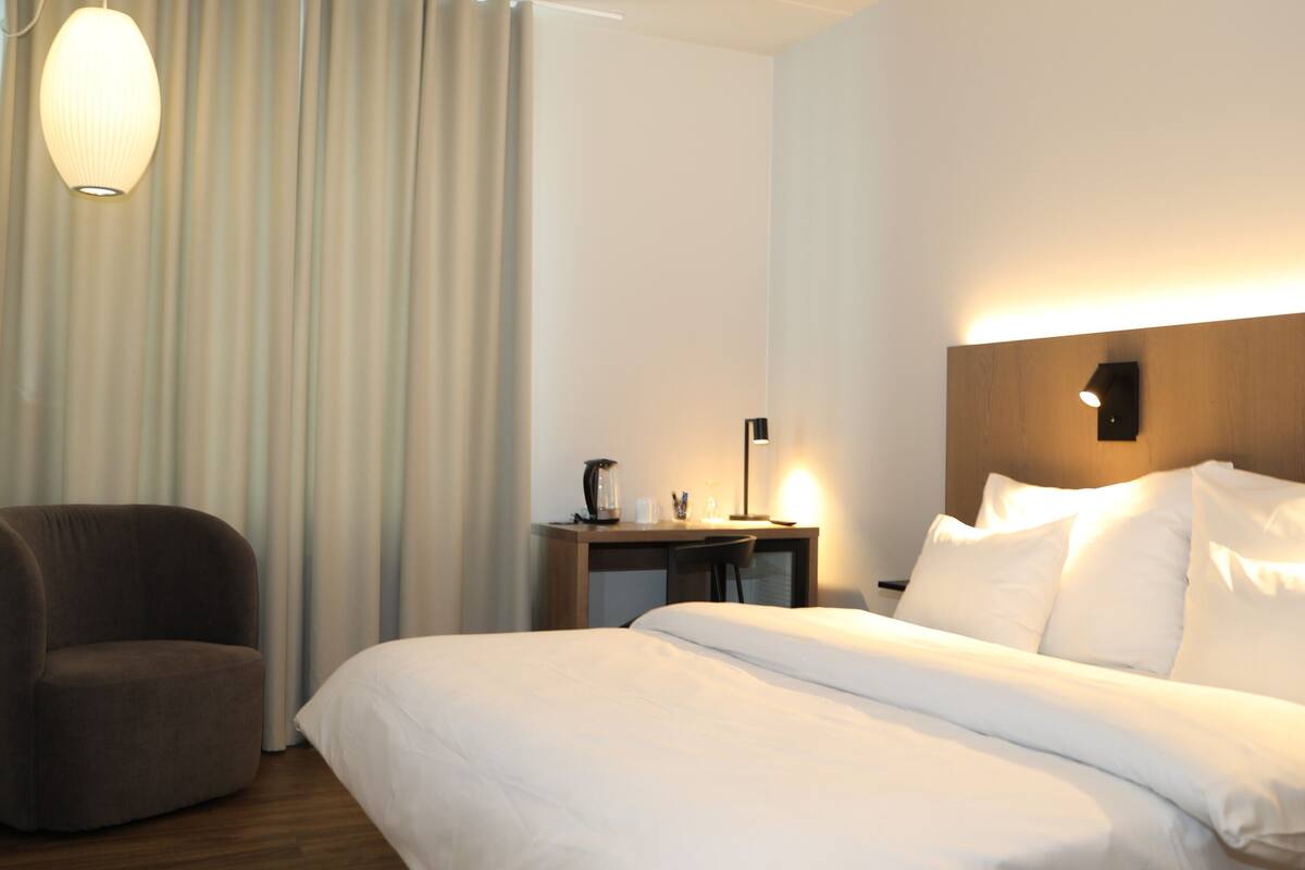 A hotel room with a bed, a chair, a desk with a lamp, and a wall-mounted light.
