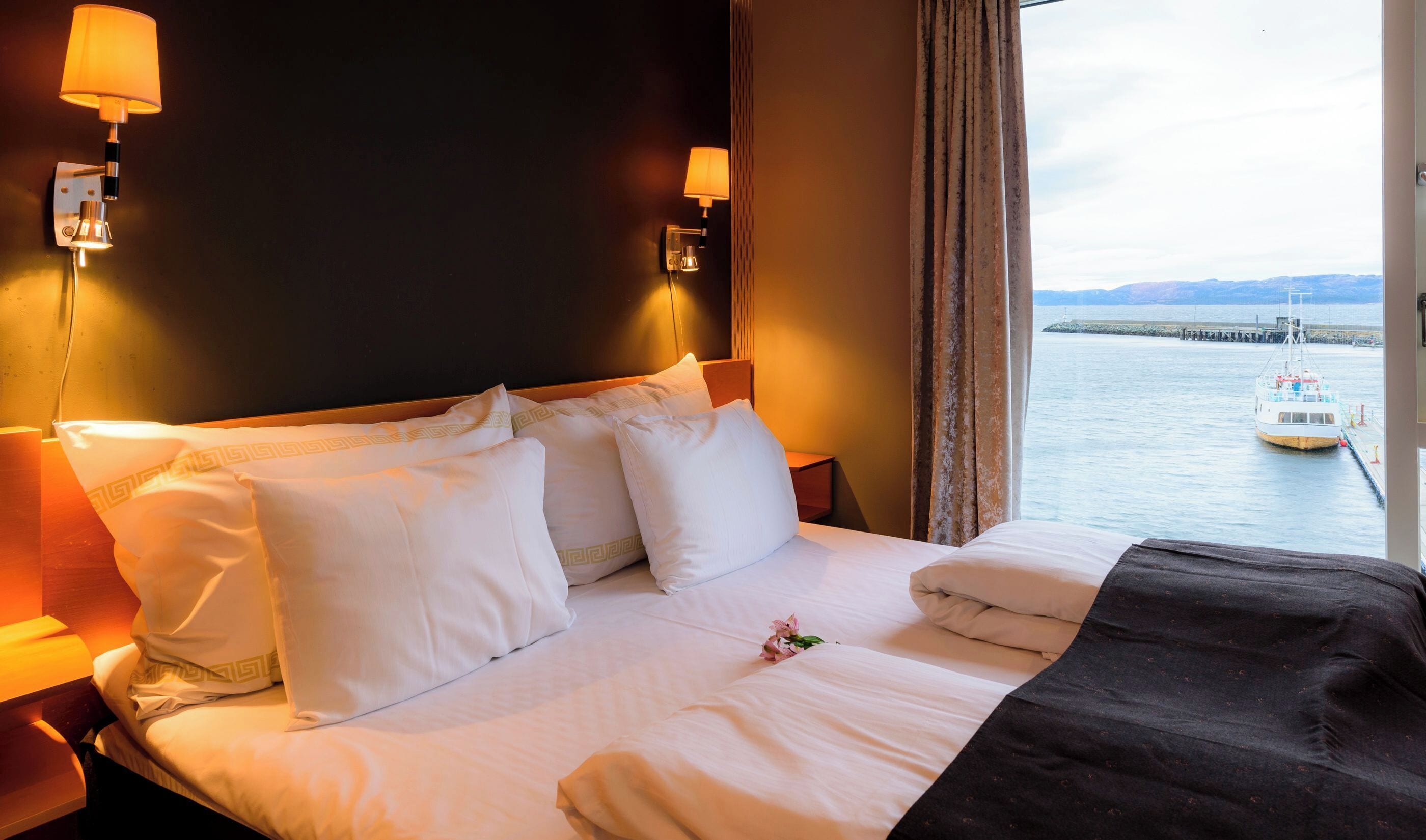 A hotel room with a bed, pillows, a nightstand, a lamp, and a view of a body of water with a boat.