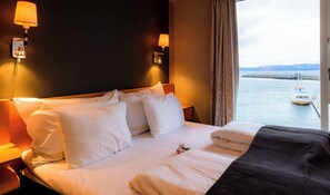 Habitación de hotel con cama, almohadas, mesita de noche, lámpara y vista a un cuerpo de agua con un barco.