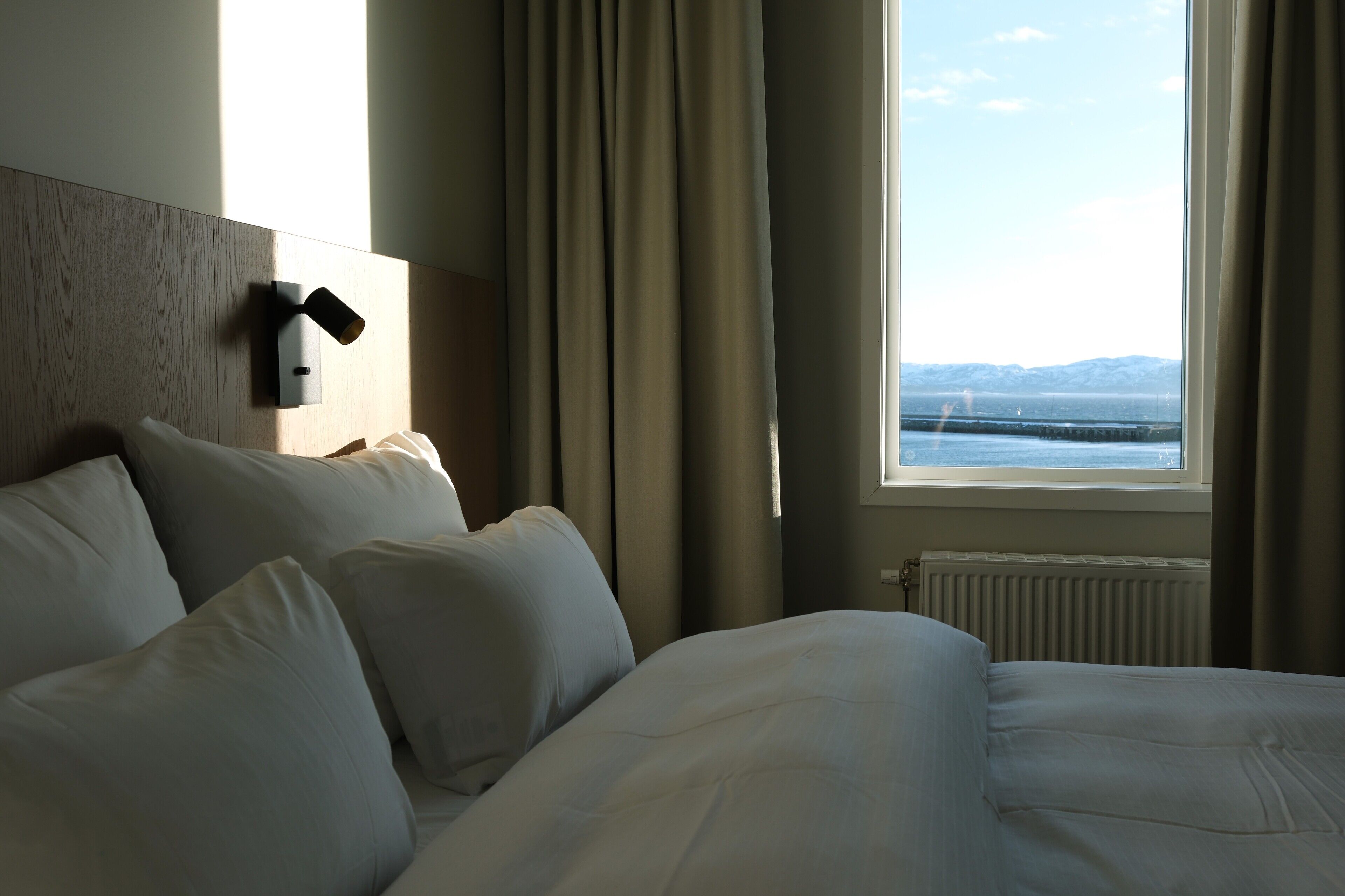 Habitación de hotel con una cama grande, cabecera, ventana con cortinas y vistas al mar y a las montañas.