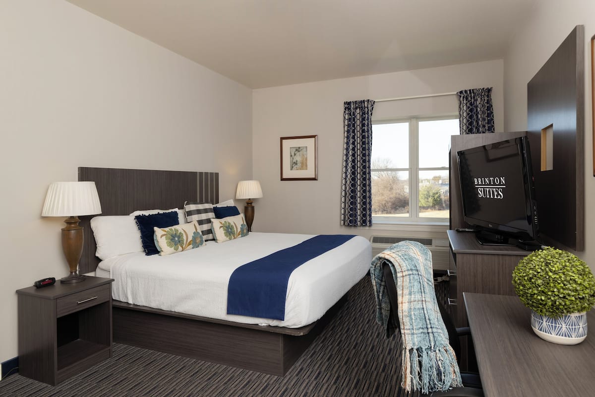 Quarto de hotel com cama, luminárias de cabeceira, televisão, escrivaninha e janela com cortinas.