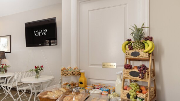 Una mesa de buffet con diversos alimentos, un televisor mostrando 'BRINTON SUITES' y un letrero que dice 'DEVOLUCIÓN DE LLAVES'.