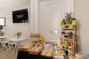 Una mesa de buffet con diversos alimentos, un televisor mostrando 'BRINTON SUITES' y un letrero que dice 'DEVOLUCIÓN DE LLAVES'.