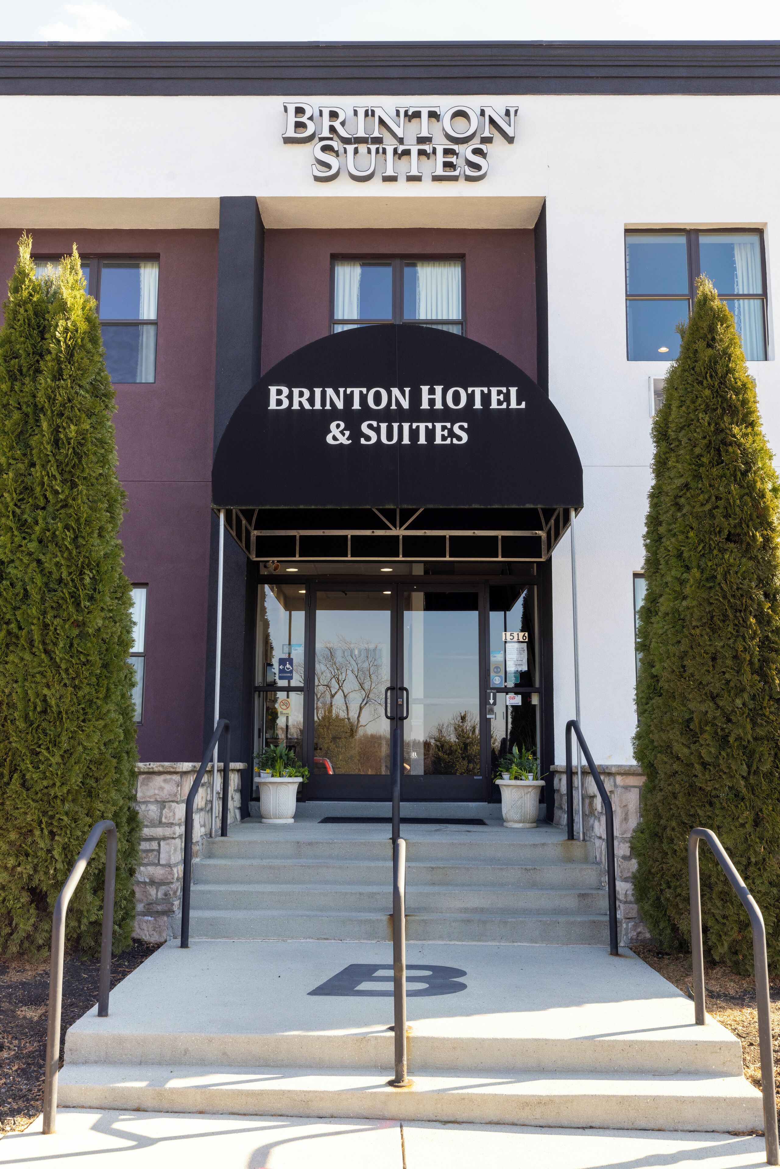 The entrance to Brinton Hotel & Suites with a clear sign, a set of stairs, and two potted plants on either side.