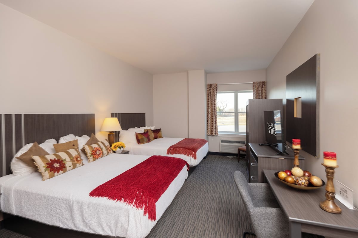Quarto de hotel com duas camas, televisão, secretária com uma tigela de fruta e janela com cortinas.