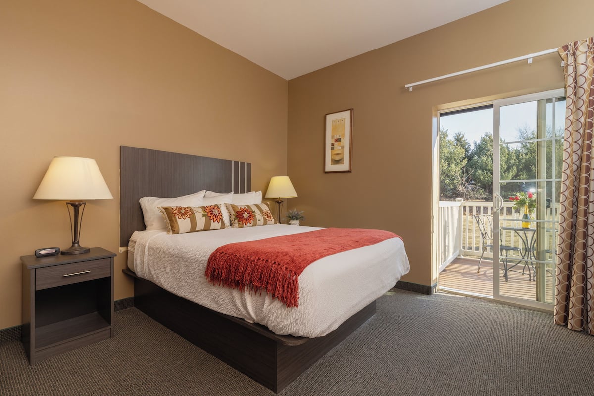 Quarto de hotel com uma cama grande, duas luminárias de cabeceira, uma mesinha de cabeceira pequena, um quadro na parede e uma varanda com mesa e cadeiras.