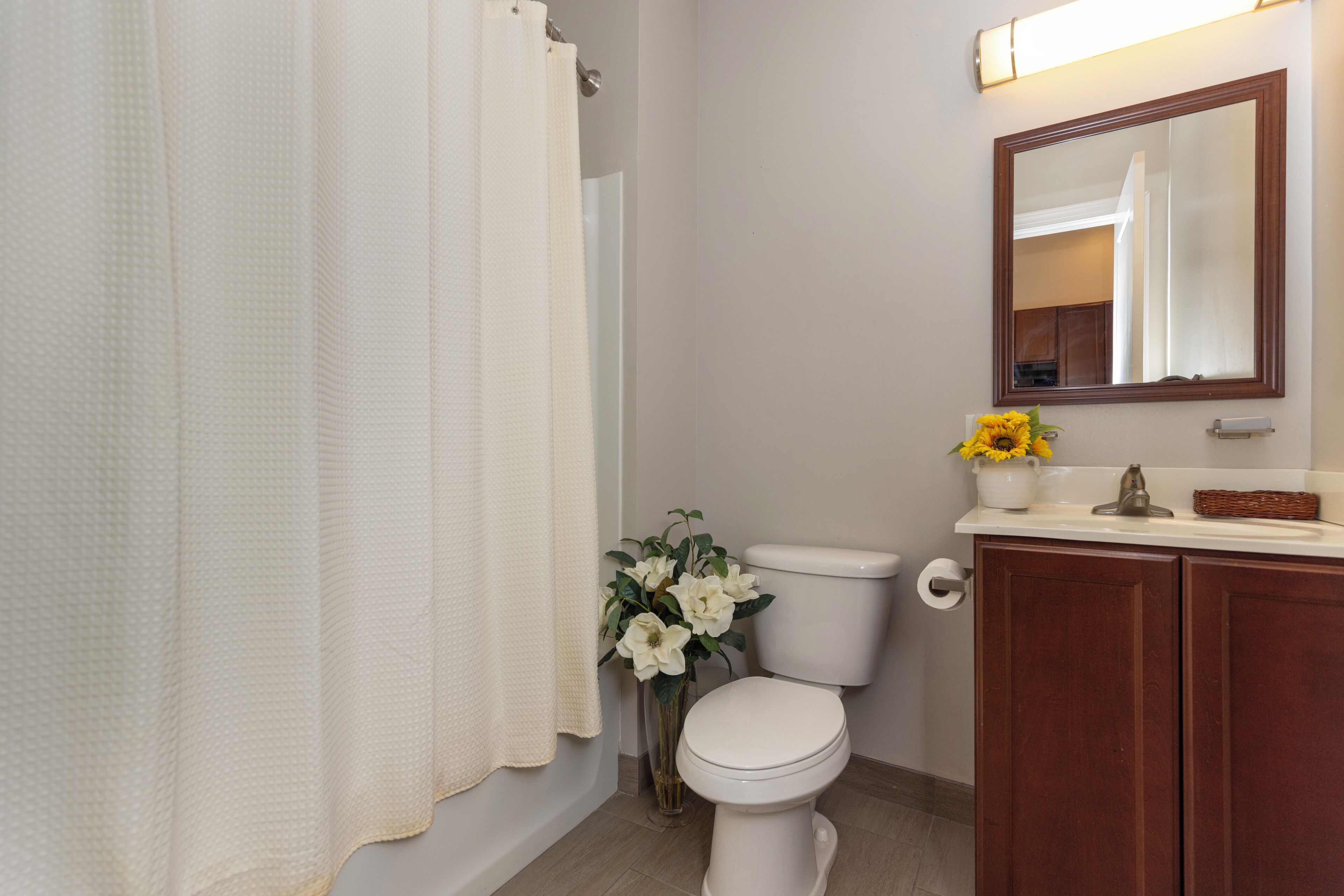A bathroom with a toilet, a sink with a mirror, a vase with flowers, and a shower curtain.
