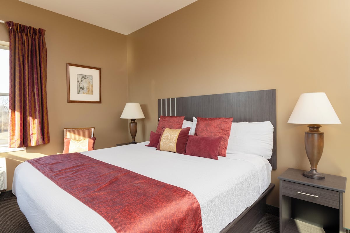Quarto de hotel com uma cama grande, duas mesas de cabeceira com luminárias, uma cadeira, um quadro emoldurado na parede e uma janela com cortinas.