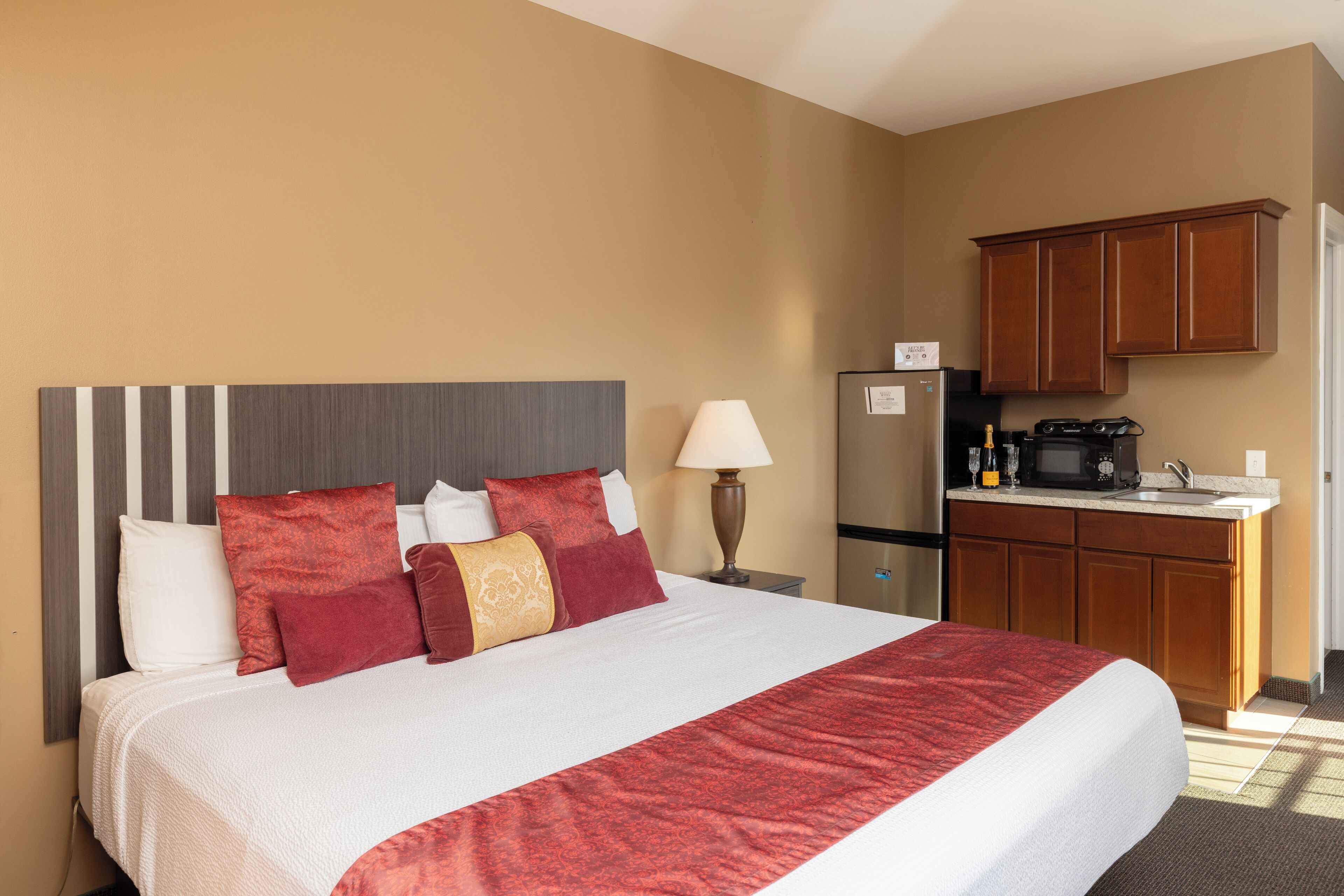 A hotel room with a large bed, a nightstand, a lamp, a refrigerator, and a kitchenette with wooden cabinets.