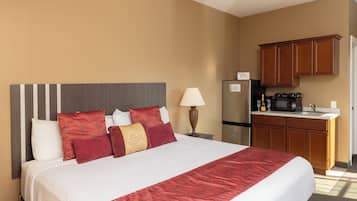 A hotel room with a large bed, a nightstand, a lamp, a refrigerator, and a kitchenette with wooden cabinets.