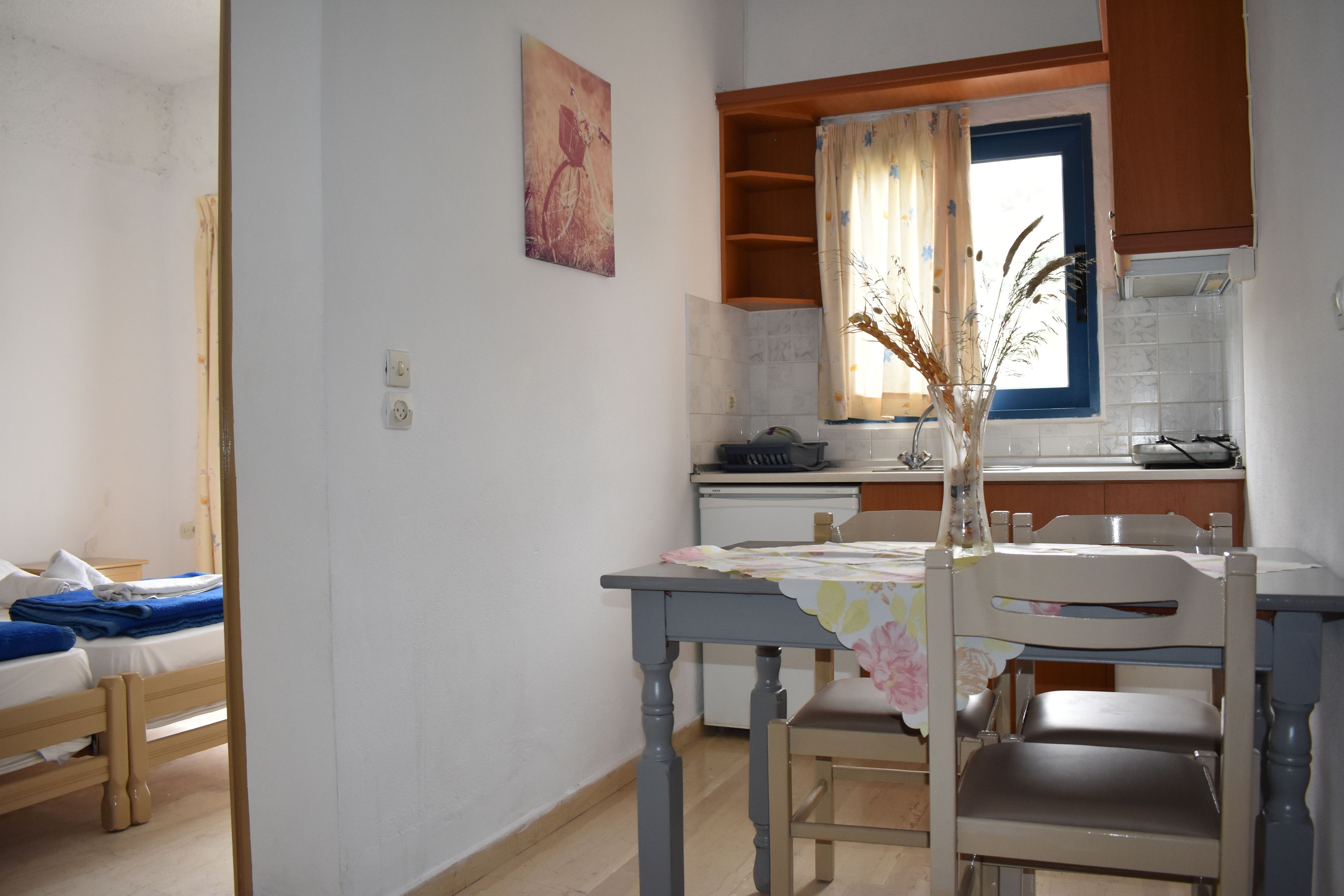 A small kitchen with a window, a dining table with chairs, and a bed in the adjacent room.