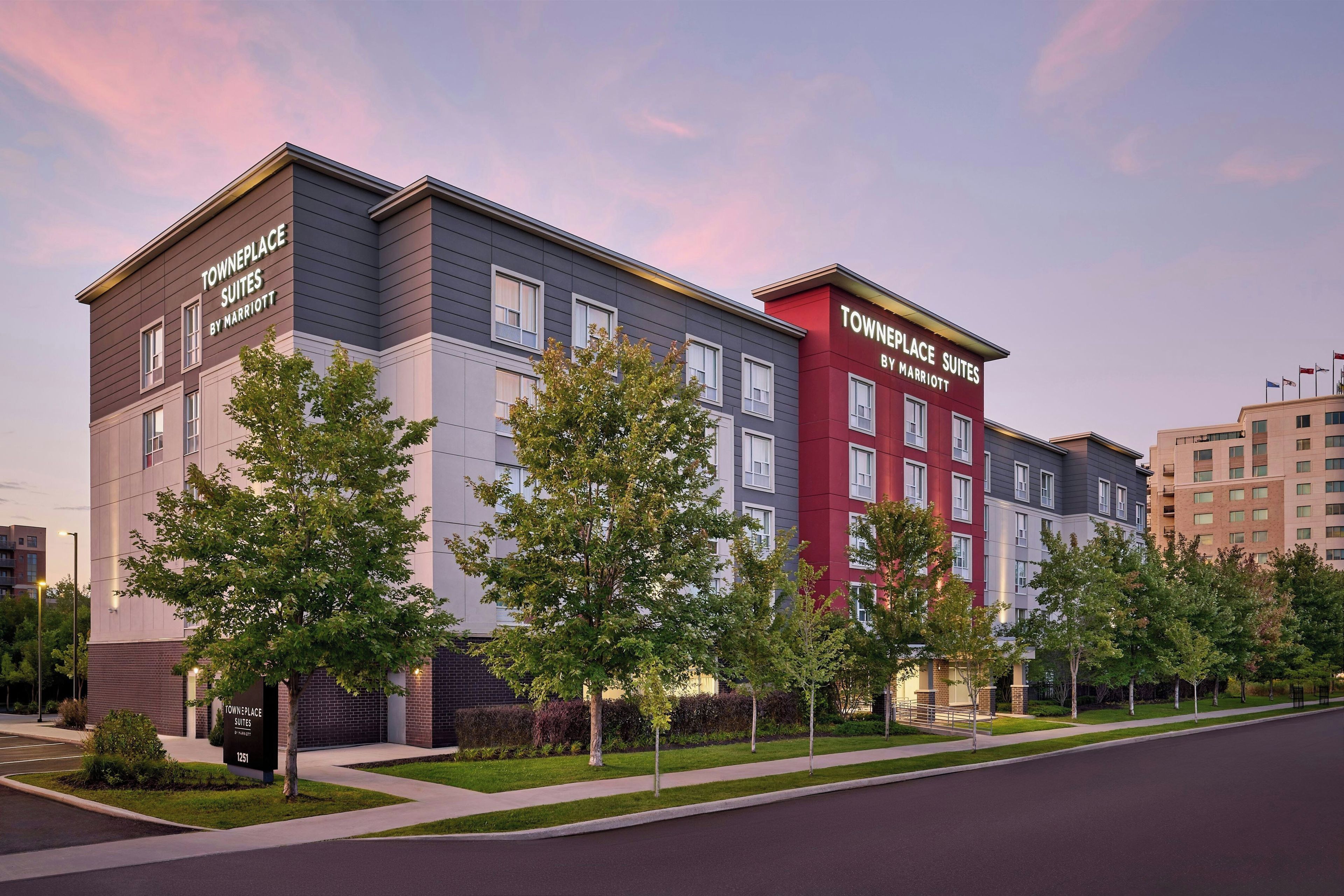 A multi-story hotel with a modern design, featuring a mix of grey and red facades, and a well-maintained landscaped front yard with young trees.